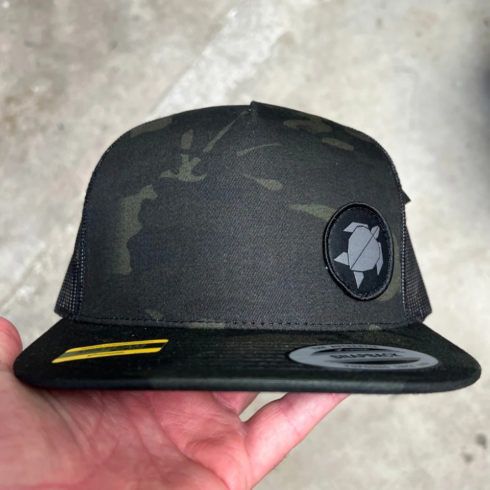 black Multicam Yupoong 6006MC mesh-back flat bill hat with small round woven turtle patch